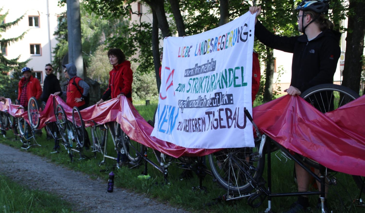 Sechs Demonstranten mit einem Banner und umgedrehten Fahrrädern, die mit roten Stoffbahnen bedeckt sind. Auf dem Banner steht: "Liebe Landesregierung, wenn ihr meint, JA zum Strukturwandel sagt endlich deutlich: Nein zu weiteren Tagebauen".