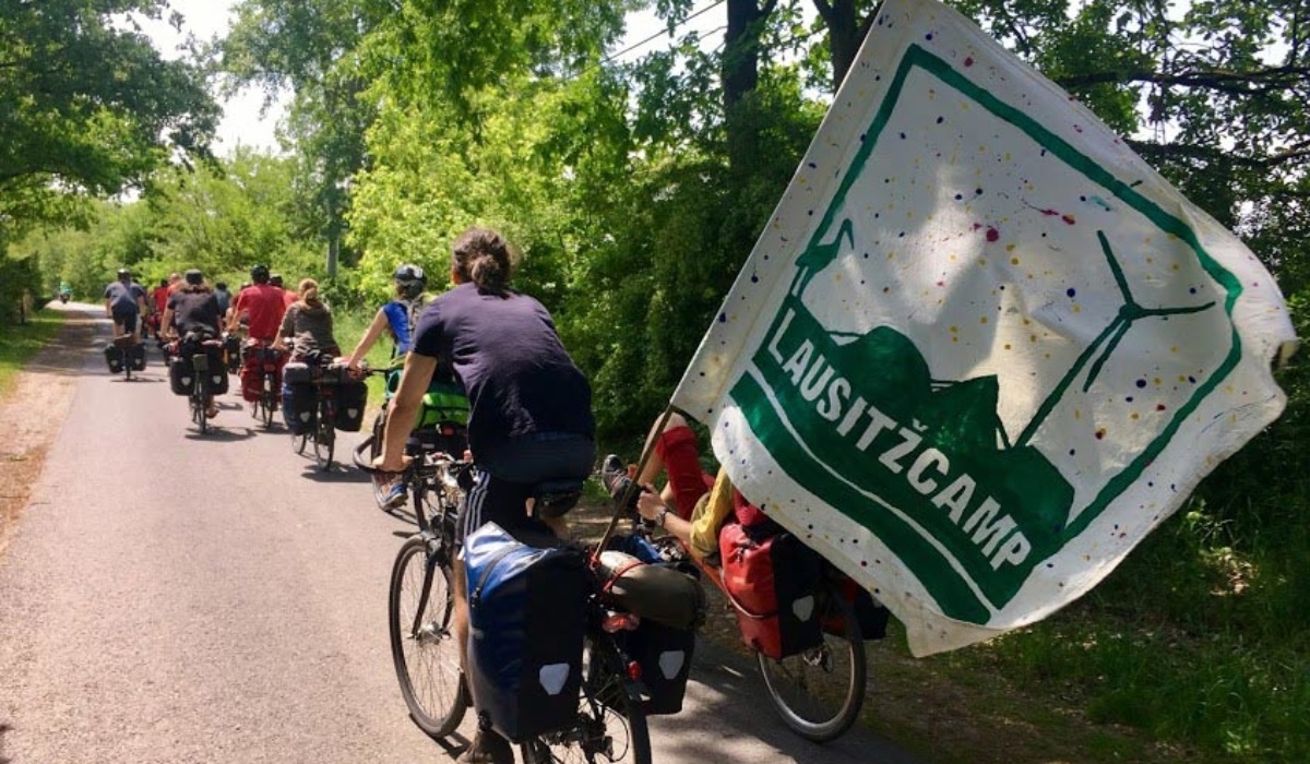 Radfahrer auf einem Asphaltweg. Im Hintergrund Bäume. Im Vordergrund eine große Fahne mit der Aufschrift: Lausitzcamp.